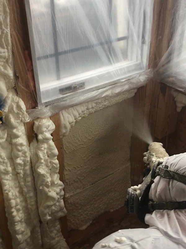Worker installing spray foam insulation in a tiny home wall beneath a window.