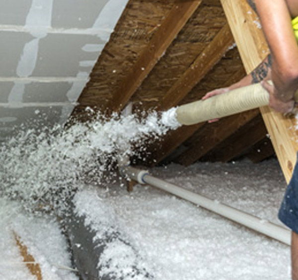 Technician spraying loose-fill insulation into attic with exposed rafters and ducting