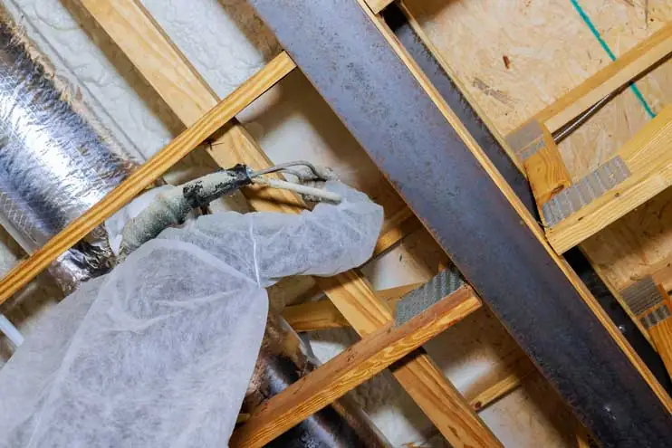 A worker in protective gear applies spray foam insulation between attic rafters to improve energy efficiency and air sealing.