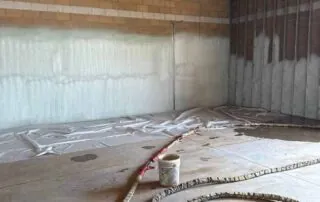 Wide shot of a commercial building interior showing newly applied closed-cell spray foam insulation on concrete block walls and exposed wood joists.