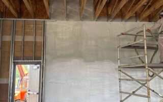 Technician applying closed-cell spray foam insulation to masonry walls inside a commercial building using a spray hose from scaffolding.