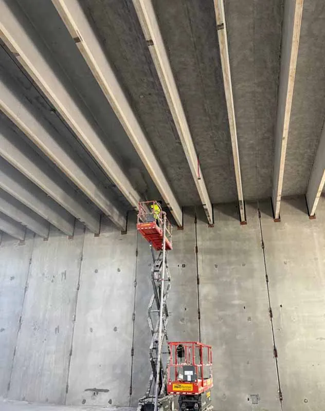 Technician applying light gray K-13 spray-on acoustic insulation to a concrete ceiling in a school gym for sound control.