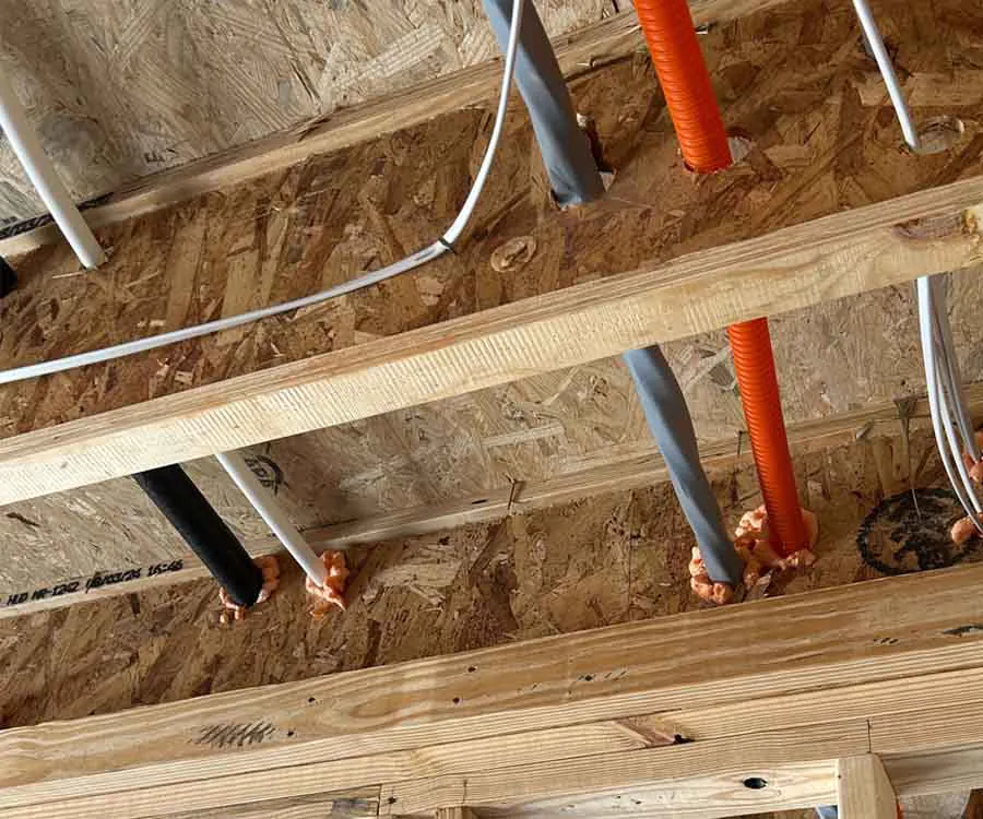 Electrical wires and cables run through drilled holes in a wooden ceiling, sealed with orange foam for insulation and fireproofing