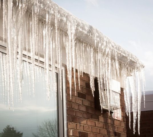 Gutters filled with an ice dam and large icicles hanging from the roof of a brick building.