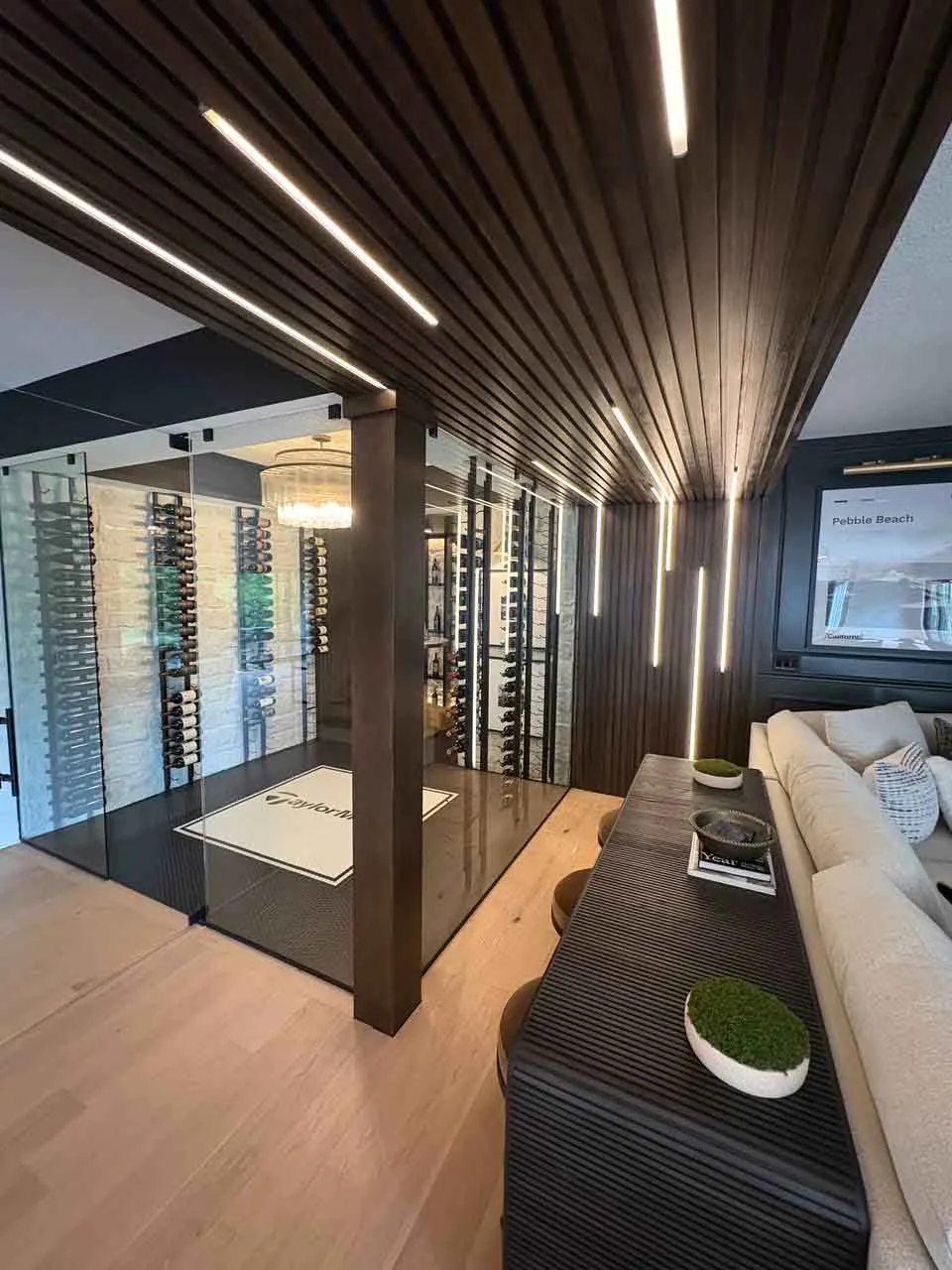 Two-sided glass wine room Interior view of a two-sided glass wine room with wall-mounted bottle racks, dark wood slatted ceiling, and vertical LED strip lighting.
