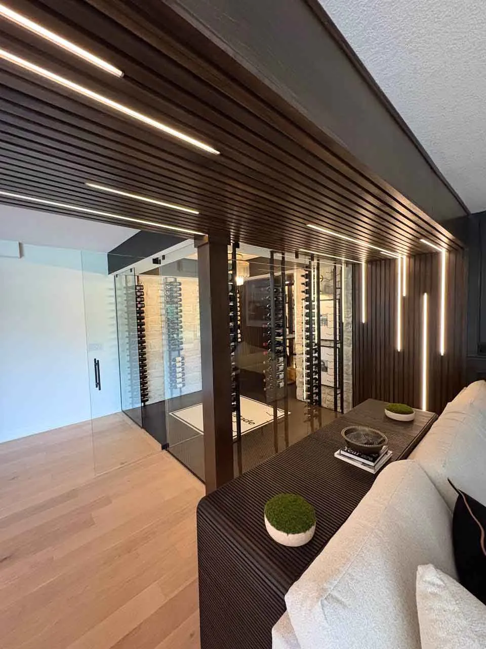 Two-sided frameless glass wine room enclosure Wide angle view of a two-sided frameless glass wine room enclosure with linear LED ceiling lighting, bottle racks, and adjacent lounge seating.