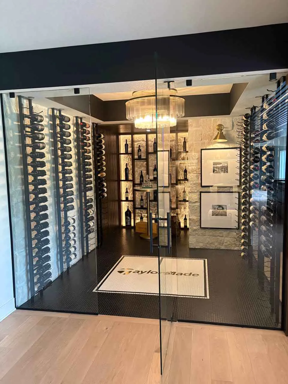 Frameless glass panels into a custom wine room Front view through frameless glass panels into a custom wine room with floor-to-ceiling bottle racks, stone accent wall, pendant chandelier, and TaylorMade logo floor mat.