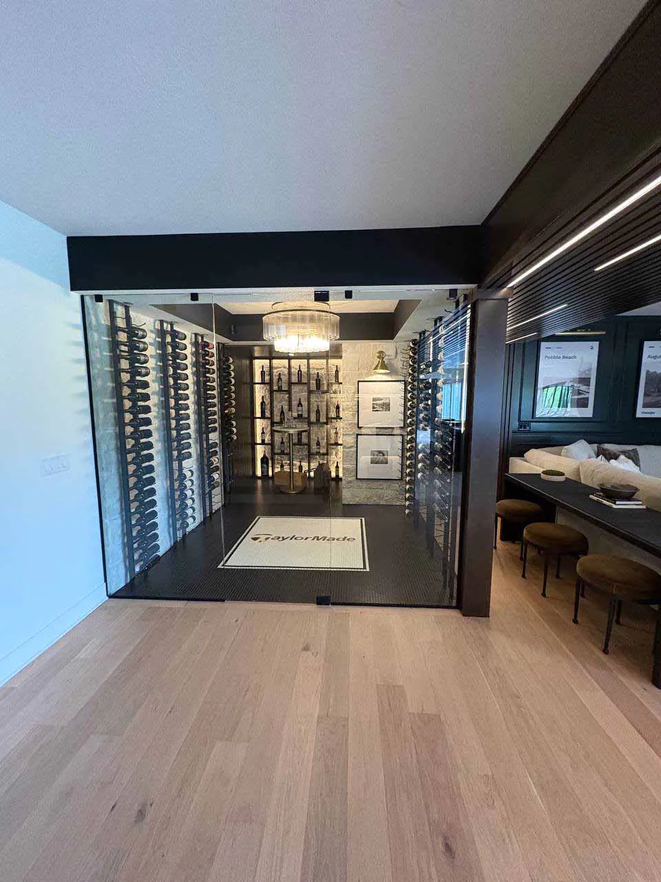 Two-sided frameless glass wine room Straight-on exterior view of a two-sided frameless glass wine room with bottle racks on both sides, stone accent wall, pendant chandelier, and TaylorMade logo floor mat.