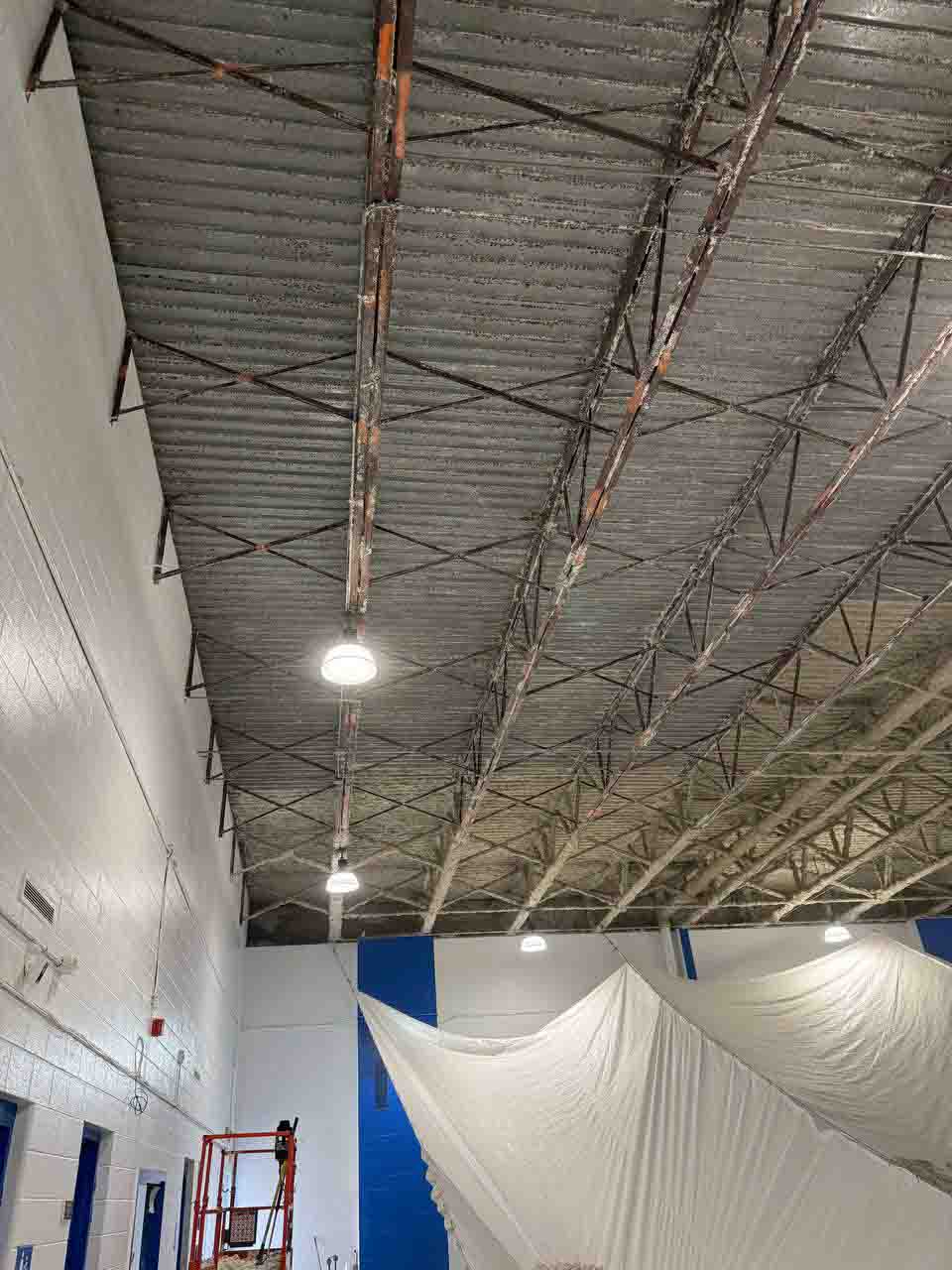 Wide view of uninsulated commercial gym ceiling with steel trusses and protective drop cloth below.