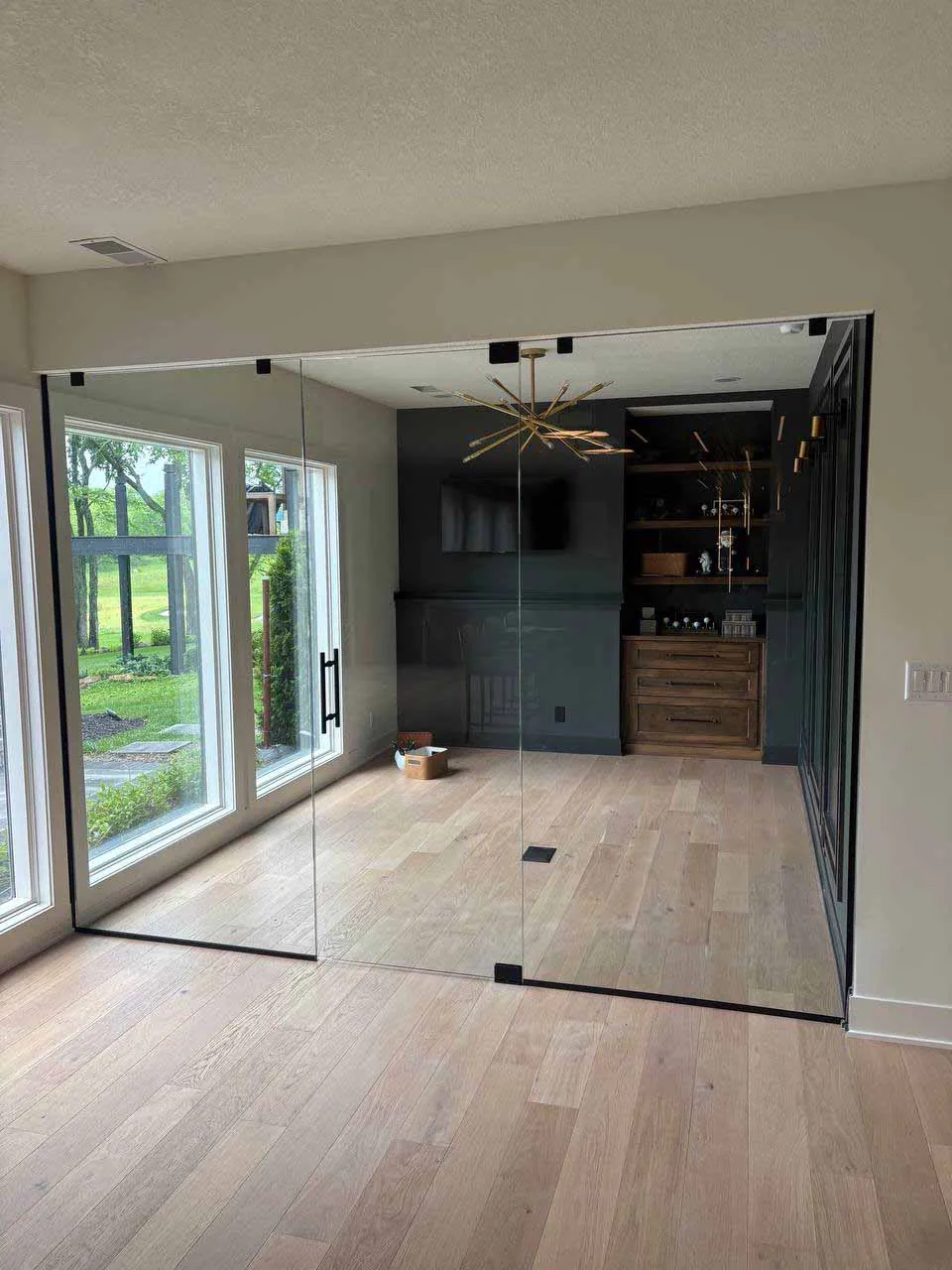Two-panel frameless glass enclosure with a single glass door creating a home office. Two-panel frameless glass enclosure with a single glass door creating a home office with large windows and hardwood floors.