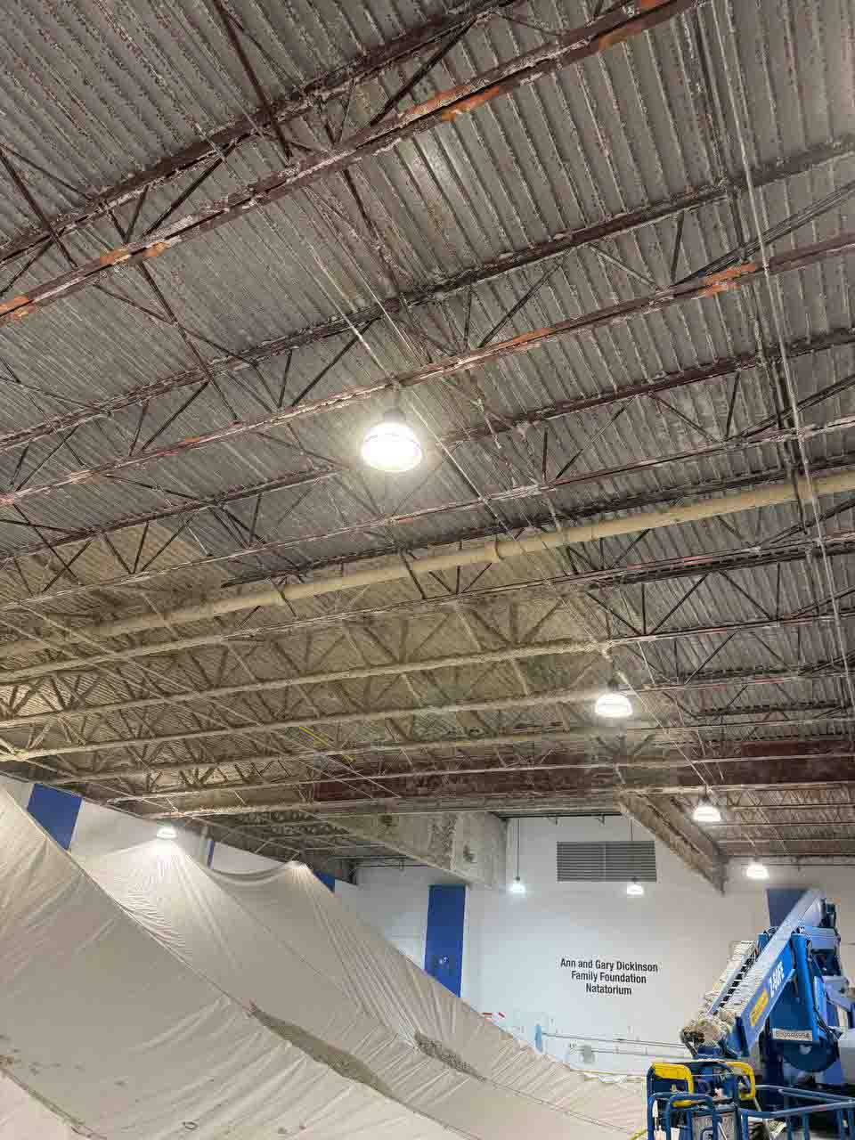 Wide view of uninsulated natatorium ceiling with steel trusses, drop cloths, and boom lift in position.