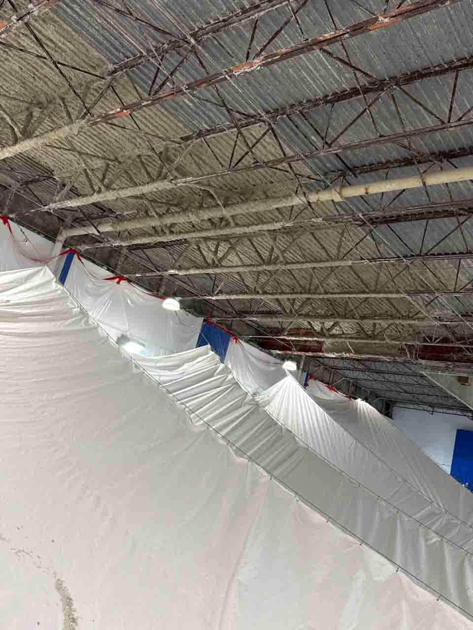 Large white containment drop cloths suspended below natatorium steel trusses during insulation project.