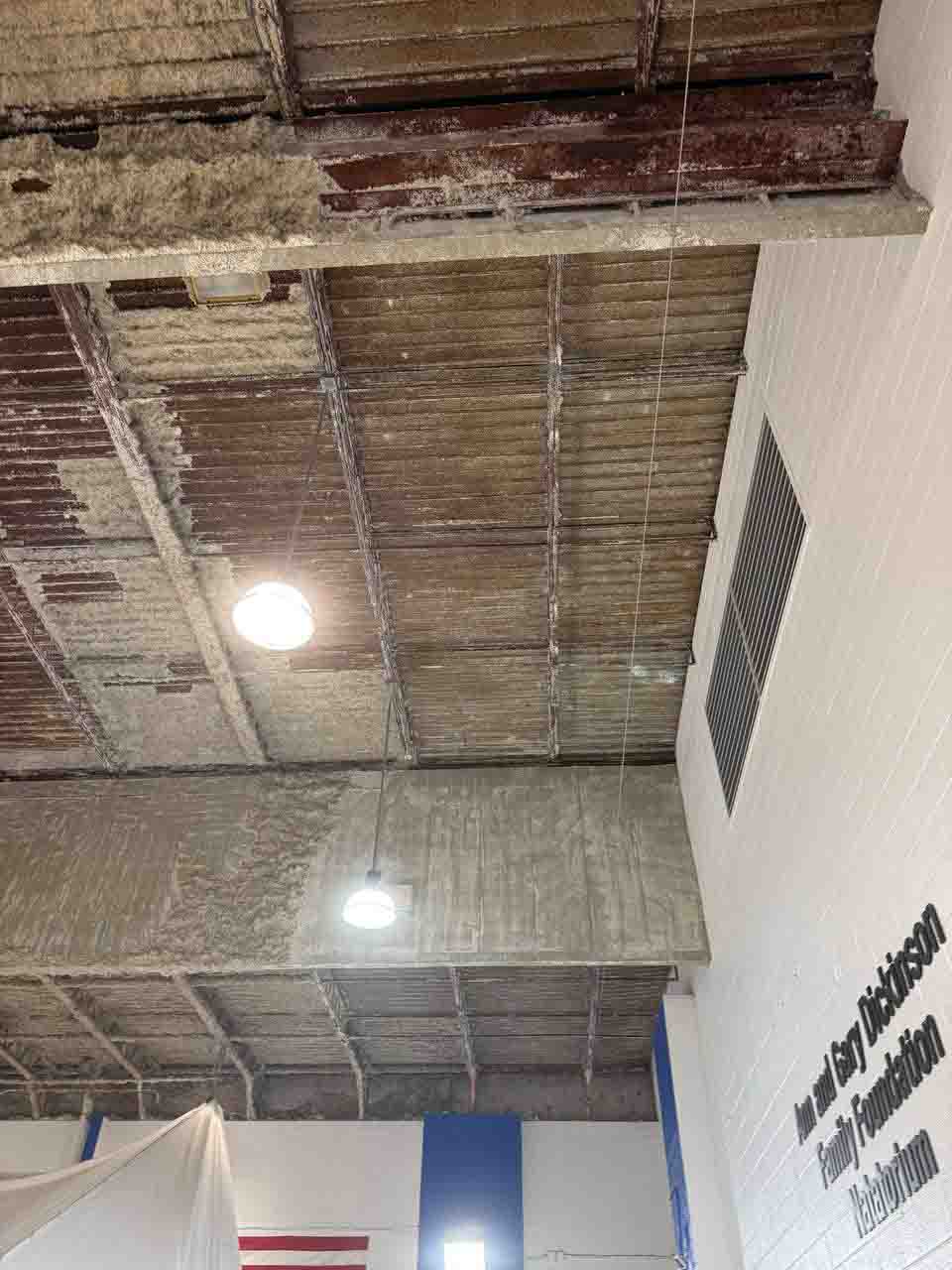 Partially spray-foamed natatorium ceiling showing insulated and bare metal deck sections side by side.