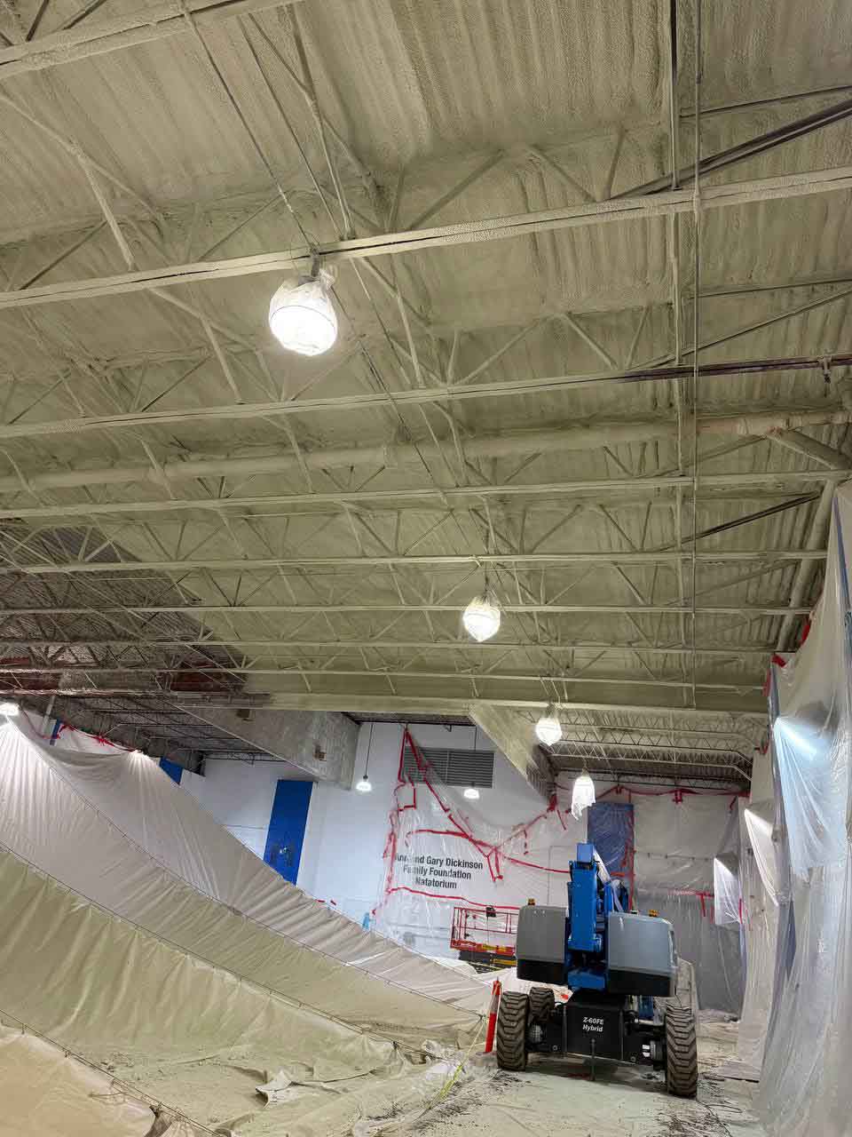 Completed spray foam insulation on natatorium ceiling with boom lift and containment tarps still in place.
