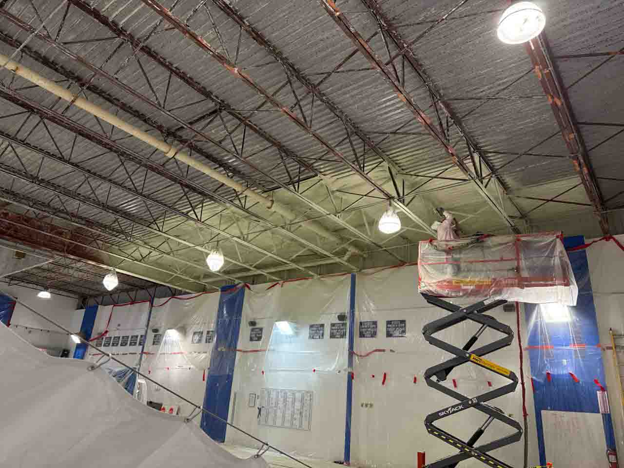 Worker on scissor lift applying spray foam insulation to commercial gym ceiling with containment barriers below.