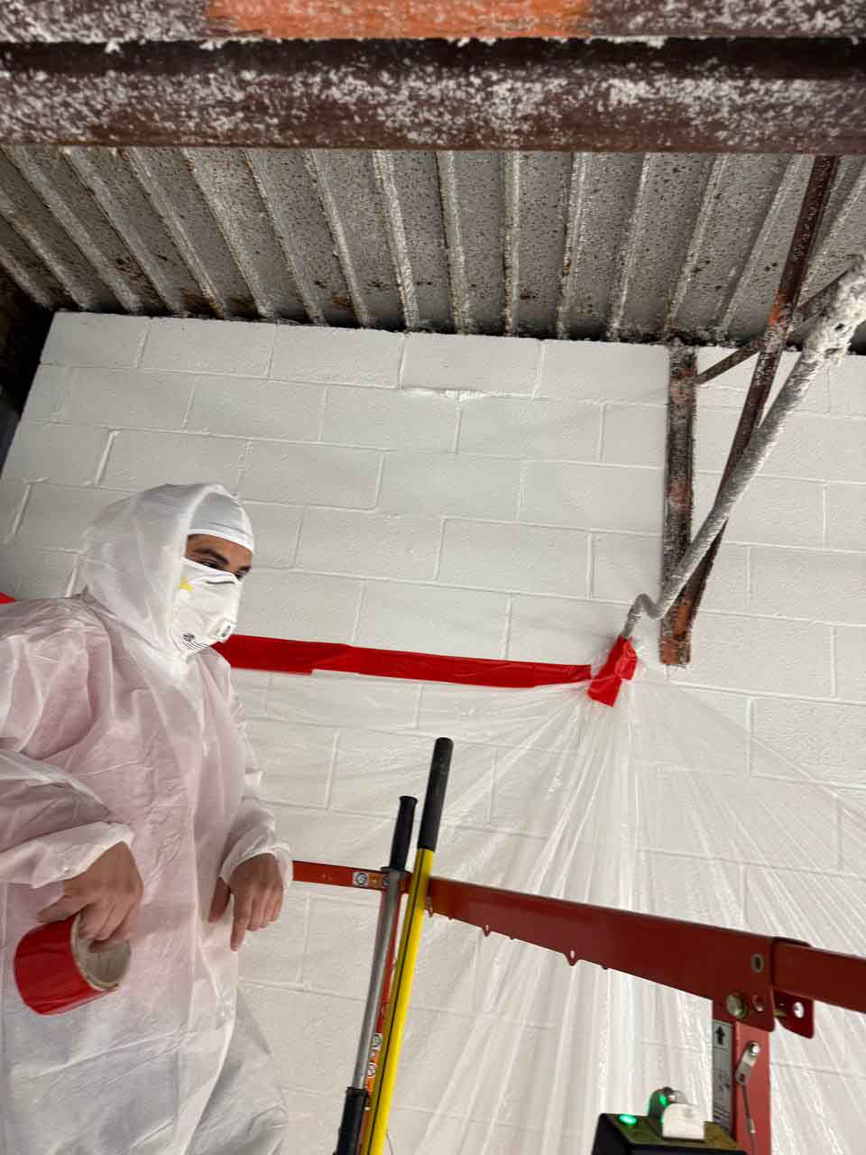 Worker in protective suit and respirator masking walls with plastic sheeting before spray foam application.