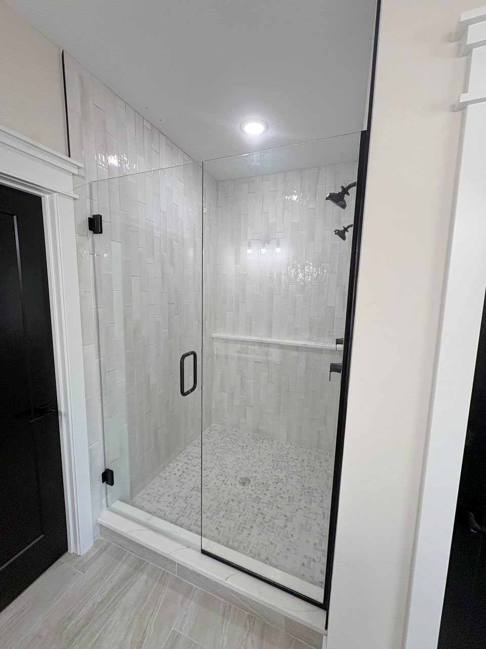 Frameless glass shower door and panel with matte black hinges and handle, enclosing a white vertical-tile shower in Kansas City.